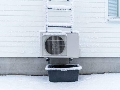 A frozen heat pump on the side of a house with the ground covered in snow
